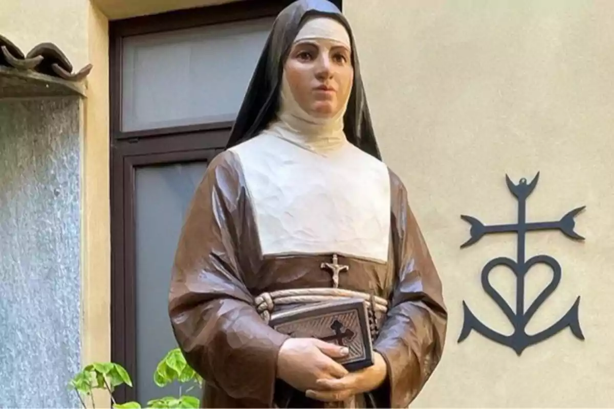 Estatua de una monja con hábito marrón y blanco sosteniendo un libro frente a una pared con un símbolo religioso en forma de cruz y corazón Estatua de una monja con hábito marrón y blanco sosteniendo un libro frente a una pared con un símbolo religioso en forma de cruz y corazón