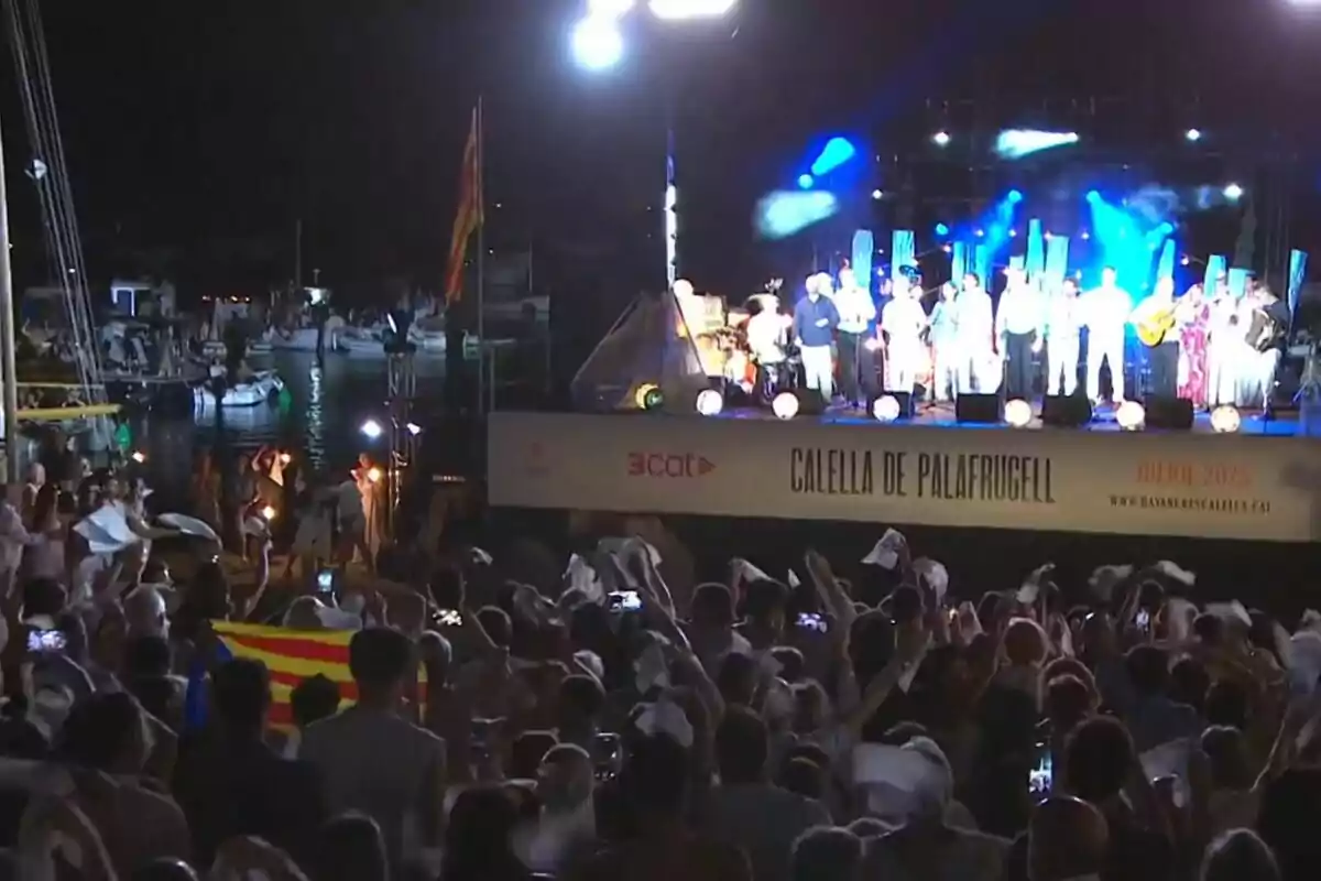 Un grup de persones gaudeix d’un concert nocturn a l’aire lliure a Calella de Palafrugell amb el públic onejant mocadors blancs i una bandera catalana visible entre la multitud. Un grup de persones gaudeix d’un concert nocturn a l’aire lliure a Calella de Palafrugell amb el públic onejant mocadors blancs i una bandera catalana visible entre la multitud.