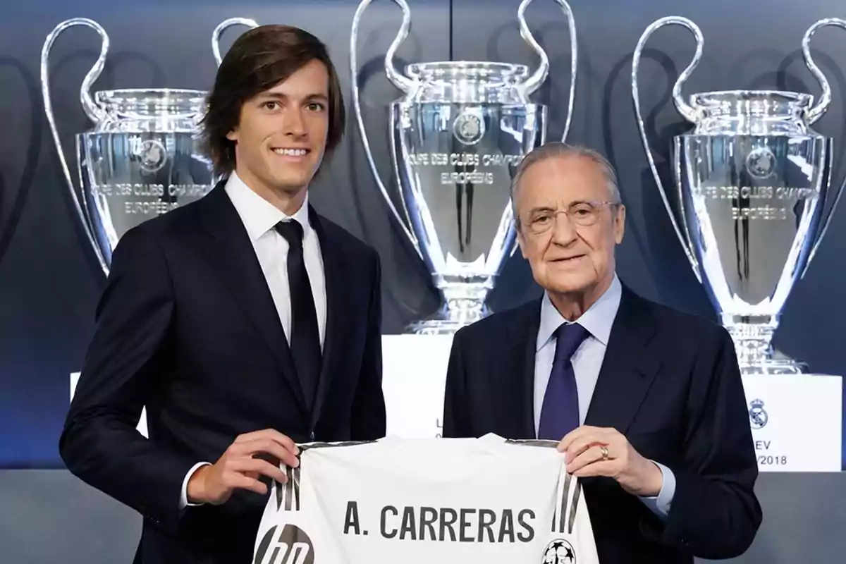Two men in suits pose smiling while holding a soccer jersey with the name A. Carreras in front of several large Champions League trophies. Two men in suits pose smiling while holding a soccer jersey with the name A. Carreras in front of several large Champions League trophies.