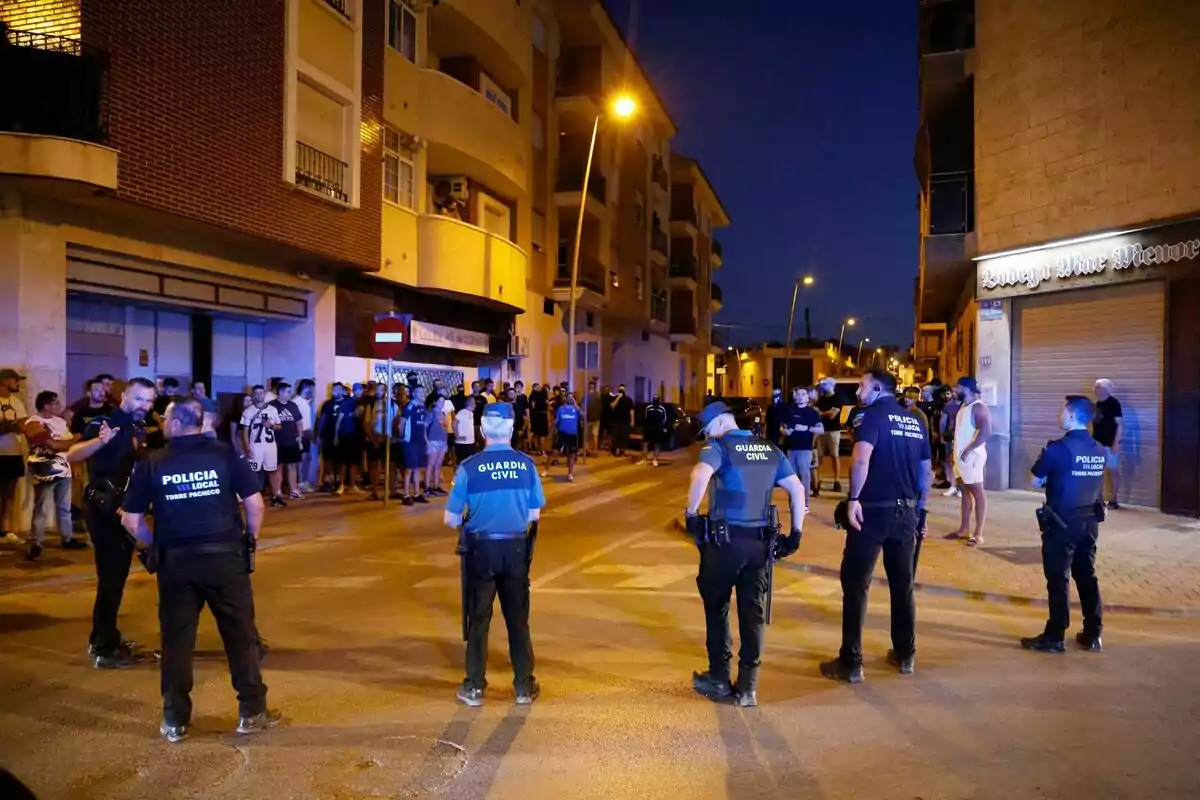 Several police officers form a barrier on a street at night while a group of people watches from the sidewalk in front of residential buildings illuminated by streetlights. Several police officers form a barrier on a street at night while a group of people watches from the sidewalk in front of residential buildings illuminated by streetlights.
