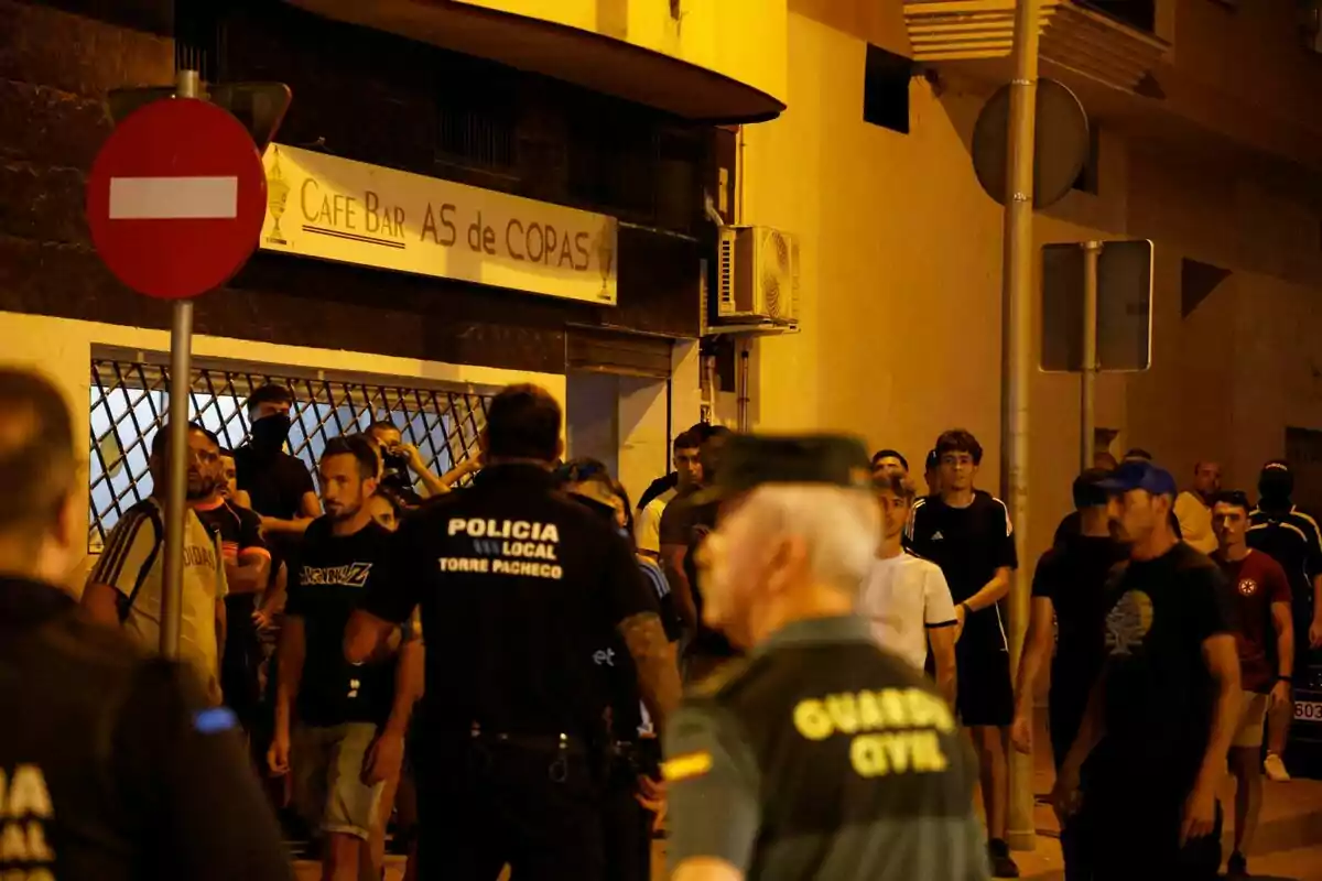 Un grup de persones i agents de policia es troben al carrer davant d’un bar durant la nit. Un grup de persones i agents de policia es troben al carrer davant d’un bar durant la nit.