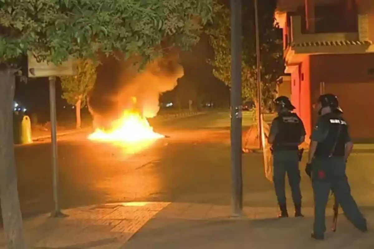 Two police officers watch a fire in the middle of a street at night.