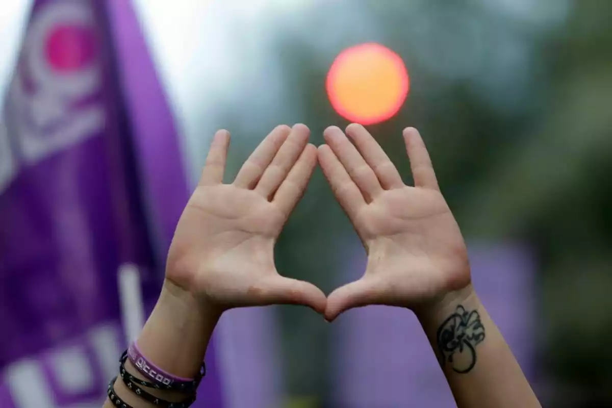 Two hands forming a triangle with their fingers in front of a blurred red light and purple flags in the background