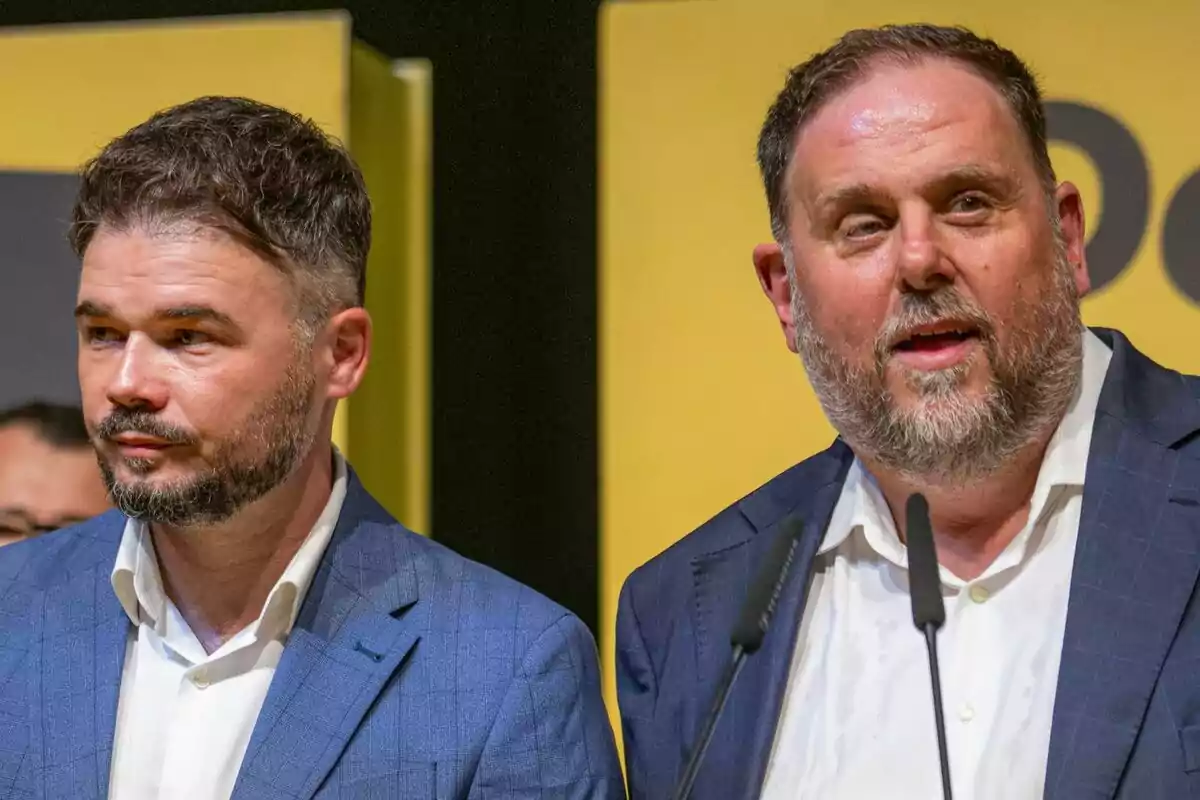 Two men with beards and blue suits pose together in front of a yellow background while one of them speaks into microphones.
