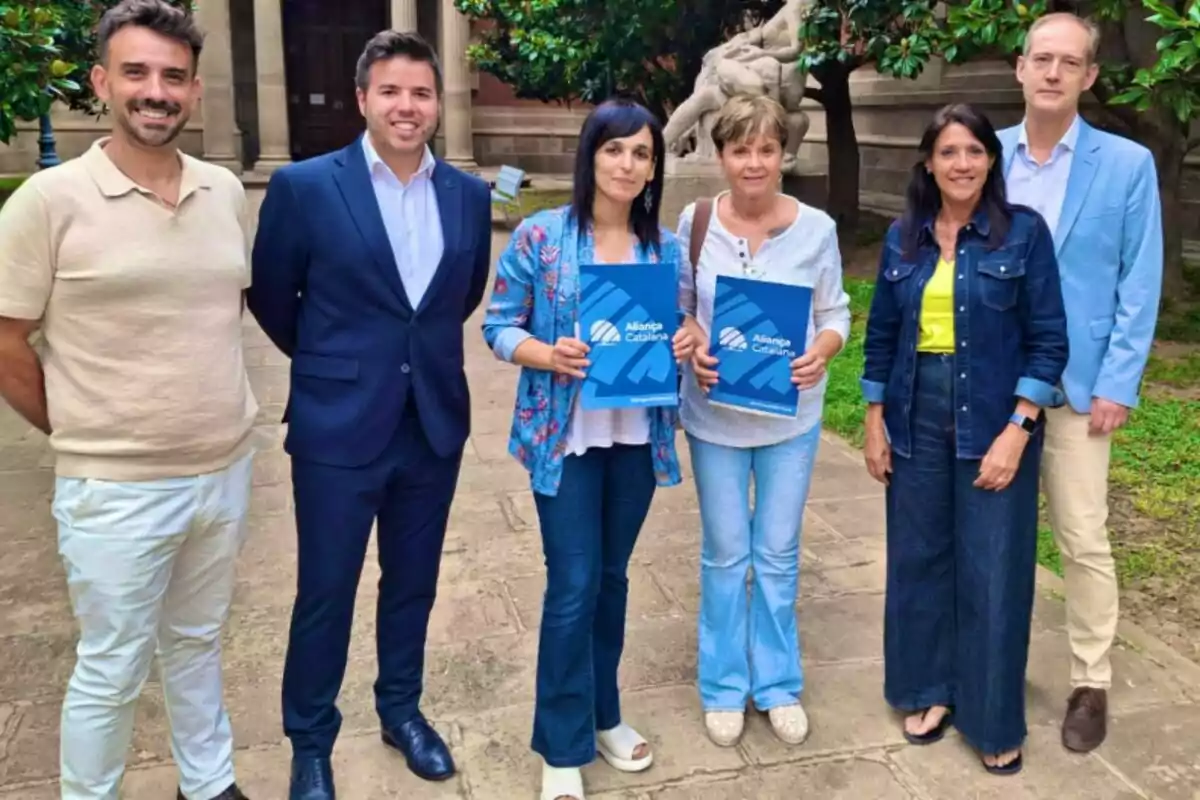 Seis personas posan al aire libre en un jardín, dos de ellas sostienen carpetas azules con el logo de Aliança Catalana.