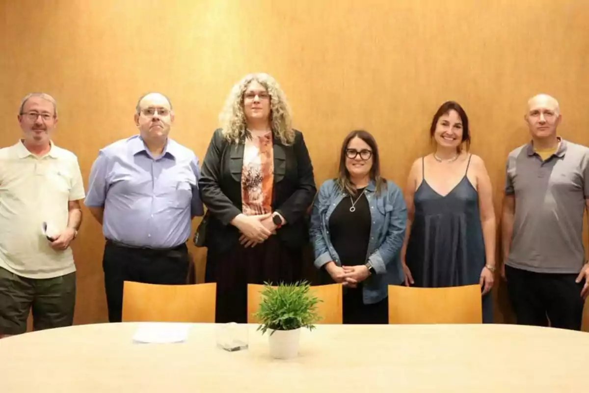 Seis personas posan de pie frente a una pared de madera detrás de una mesa con una planta en el centro