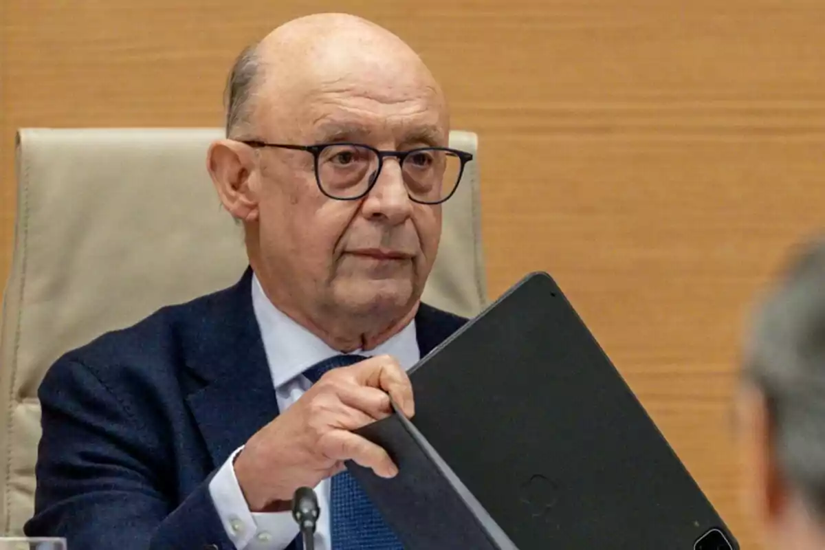 Older man with glasses and a suit holding a black tablet in a meeting room