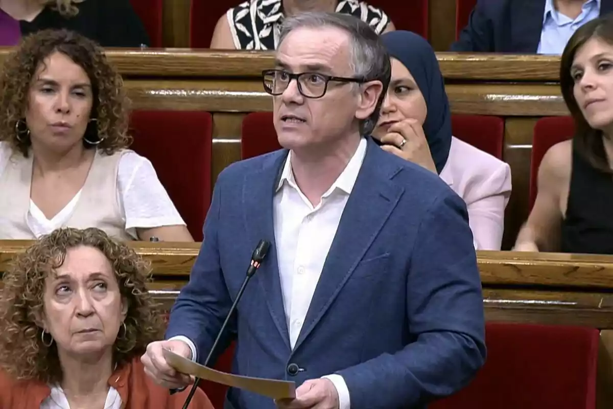 Man with glasses and a blue jacket speaking in a parliament while holding some papers, surrounded by other people seated in red benches Man with glasses and a blue jacket speaking in a parliament while holding some papers, surrounded by other people seated in red benches