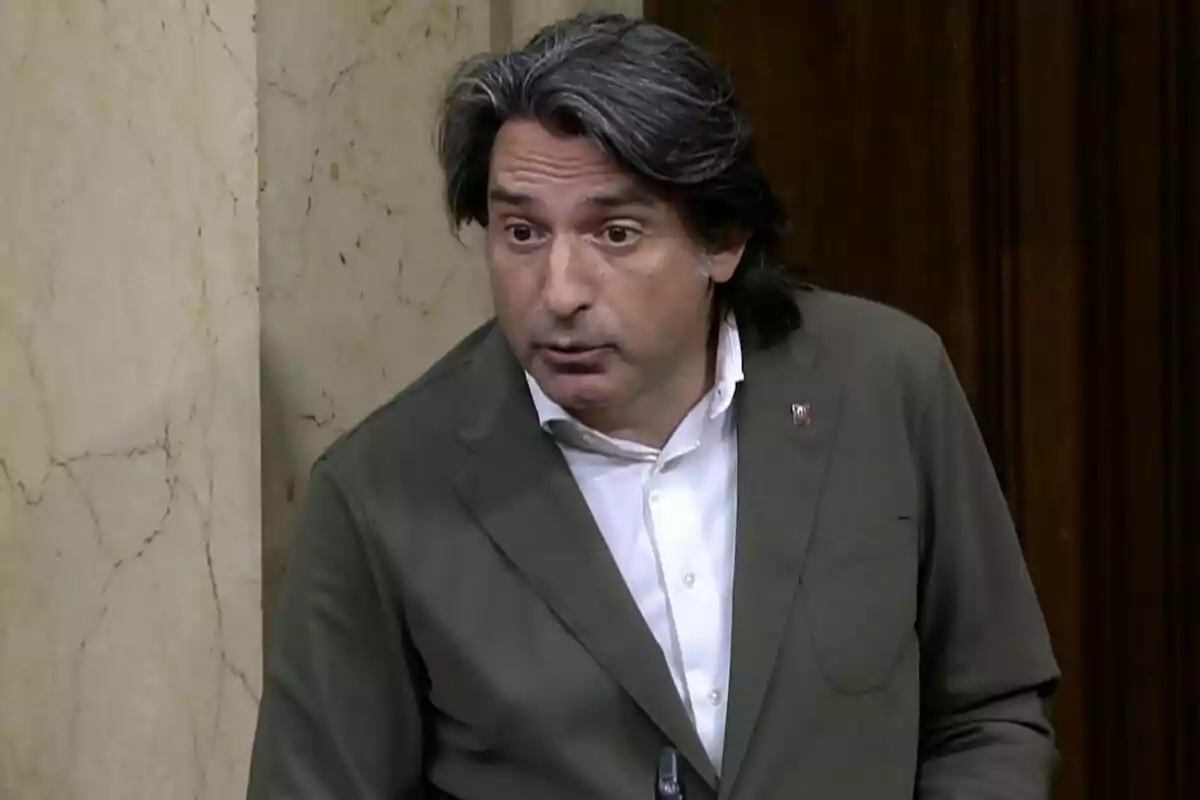 Home de cabells canosos i llargs amb jaqueta verd oliva i camisa blanca parlant en un entorn formal amb parets de marbre i fusta.