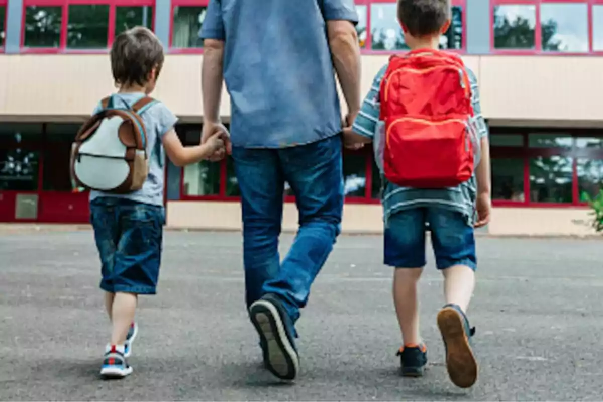 Adult walking hand in hand with two small children carrying backpacks in front of a school building