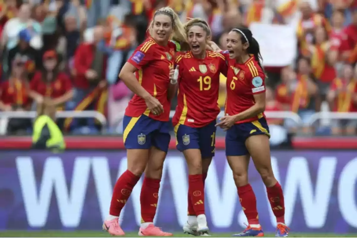 Three players from the Spanish women's national soccer team celebrate a goal on the field with expressions of joy and excitement while the crowd watches them from the stands. Three players from the Spanish women's national soccer team celebrate a goal on the field with expressions of joy and excitement while the crowd watches them from the stands.