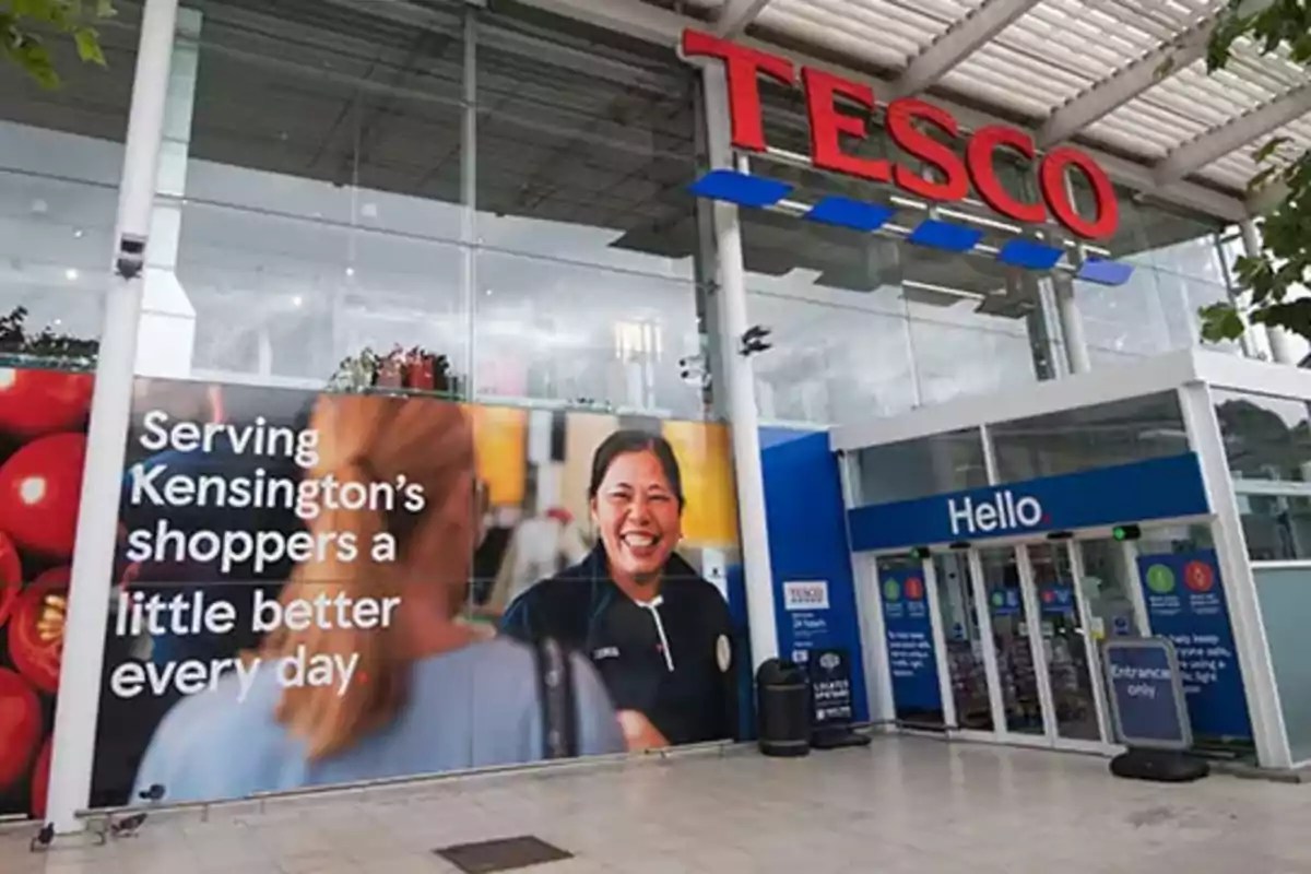 Façana d’una botiga Tesco amb un gran cartell publicitari que mostra una empleada somrient i l’entrada principal amb portes automàtiques Façana d’una botiga Tesco amb un gran cartell publicitari que mostra una empleada somrient i l’entrada principal amb portes automàtiques