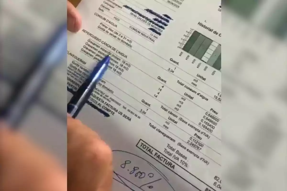 Persona assenyalant amb un bolígraf blau una factura d’aigua escrita en català on s’observen dades de consum, tarifes i un gràfic d’historial de consum a la part superior dreta