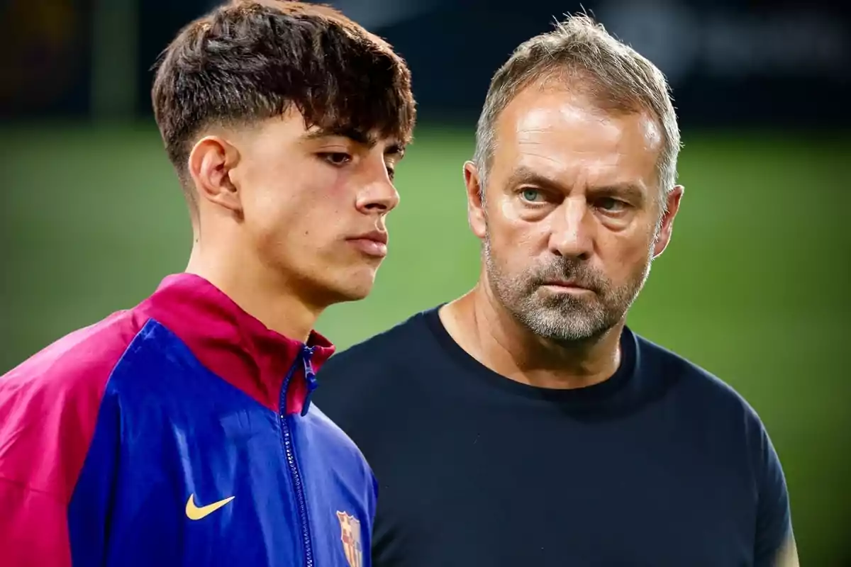 Dos personas en primer plano, una con chaqueta deportiva del Barcelona y otra con camiseta oscura, ambas con expresión seria en un entorno de campo de fútbol.