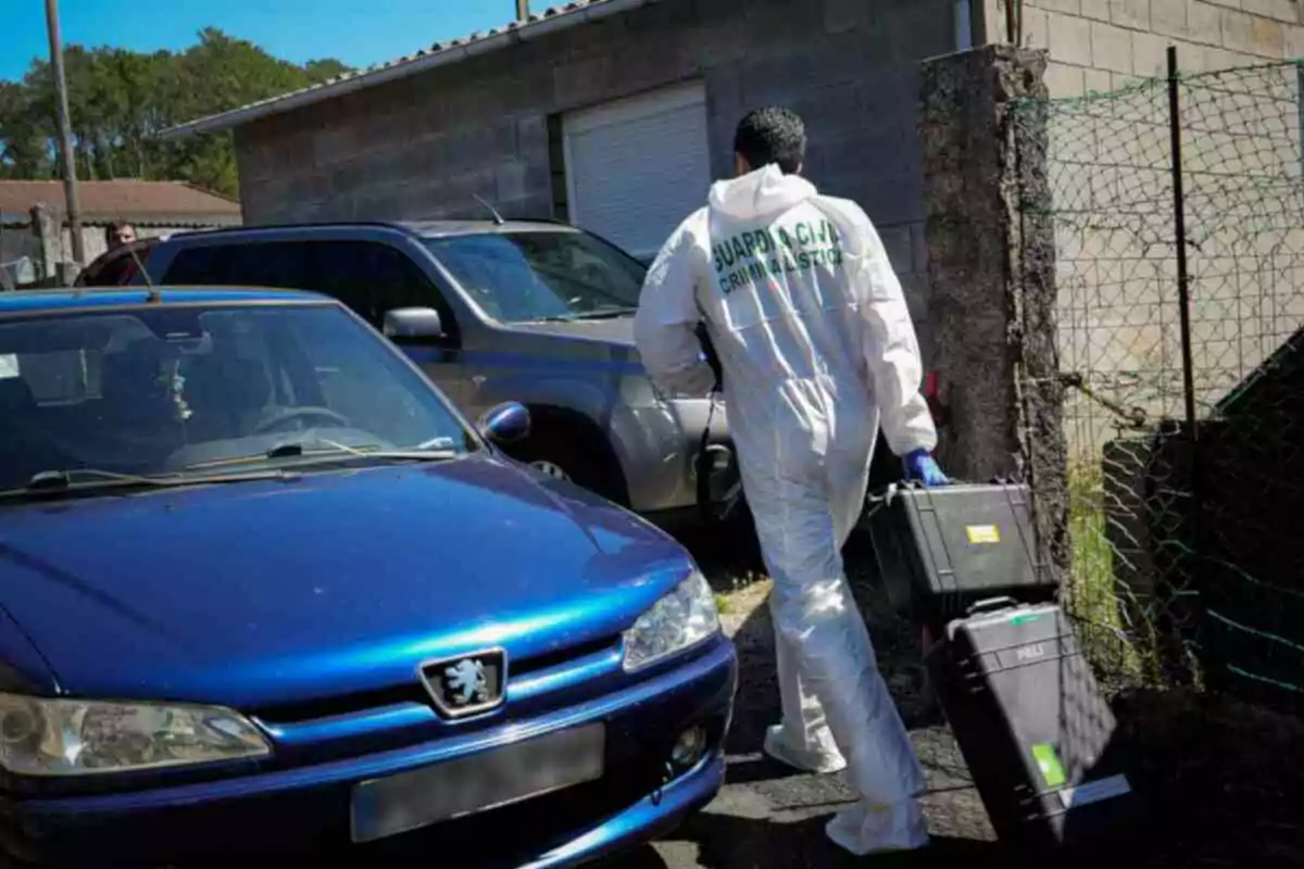 Un agent forense de la Guàrdia Civil camina al costat d’un cotxe blau portant dues maletes d’equip en una zona residencial. Un agent forense de la Guàrdia Civil camina al costat d’un cotxe blau portant dues maletes d’equip en una zona residencial.