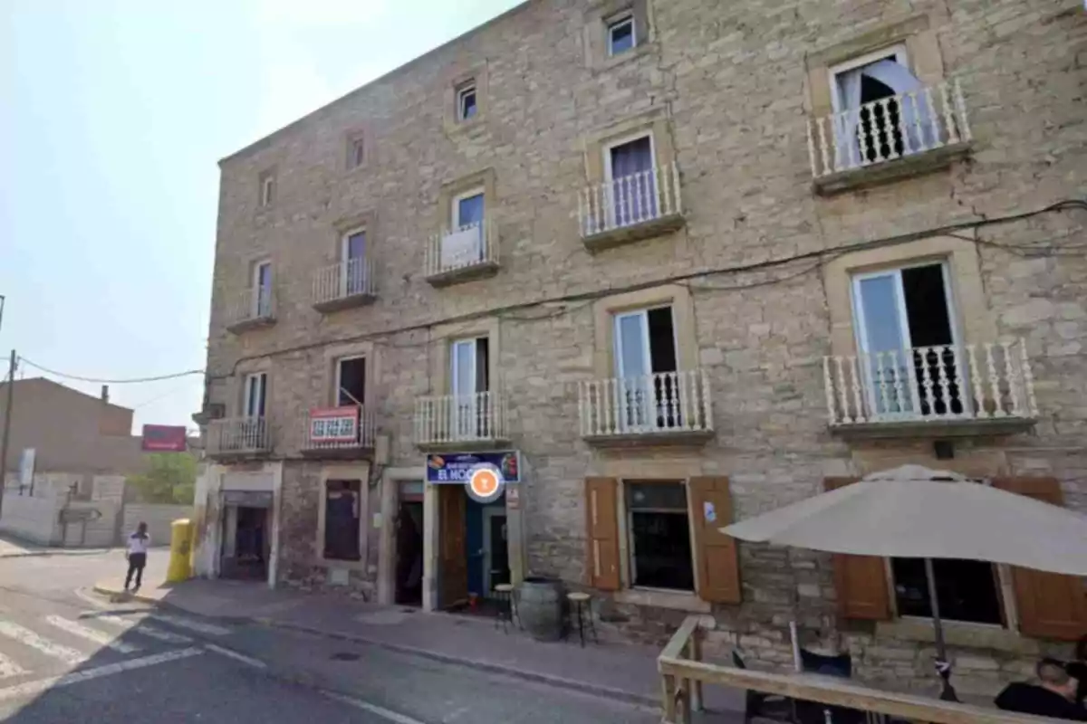 Fachada de un edificio antiguo de piedra con varias ventanas y balcones pequeños, en la planta baja hay un bar con mesas y sombrilla en la acera Fachada de un edificio antiguo de piedra con varias ventanas y balcones pequeños, en la planta baja hay un bar con mesas y sombrilla en la acera