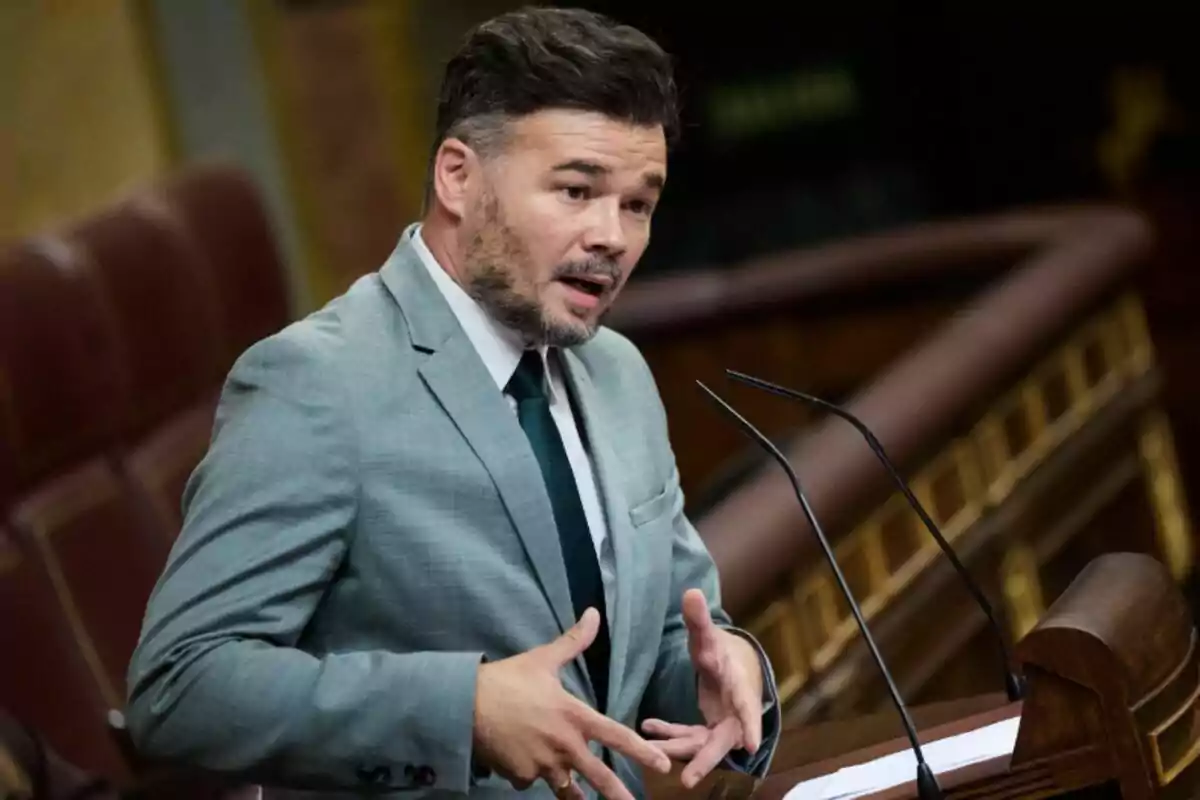 Home amb barba i cabells foscos vestit amb un vestit gris i corbata fosca parlant en un faristol en un recinte parlamentari Home amb barba i cabells foscos vestit amb un vestit gris i corbata fosca parlant en un faristol en un recinte parlamentari