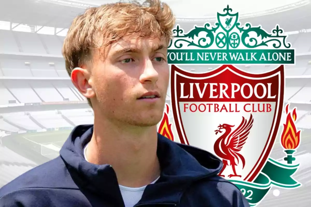 Young man in a dark jacket in front of the Liverpool Football Club crest in an empty stadium