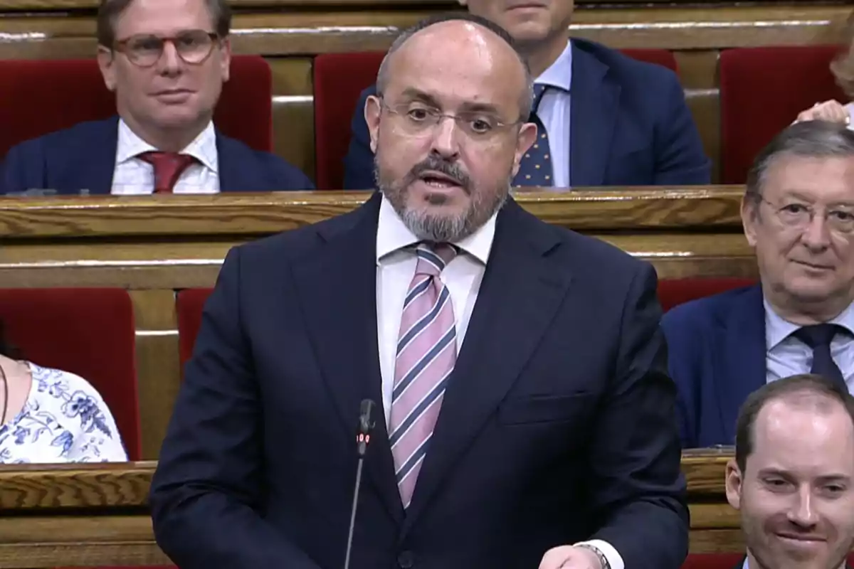 Man with a beard and glasses speaking in a parliament while other people watch him from their seats