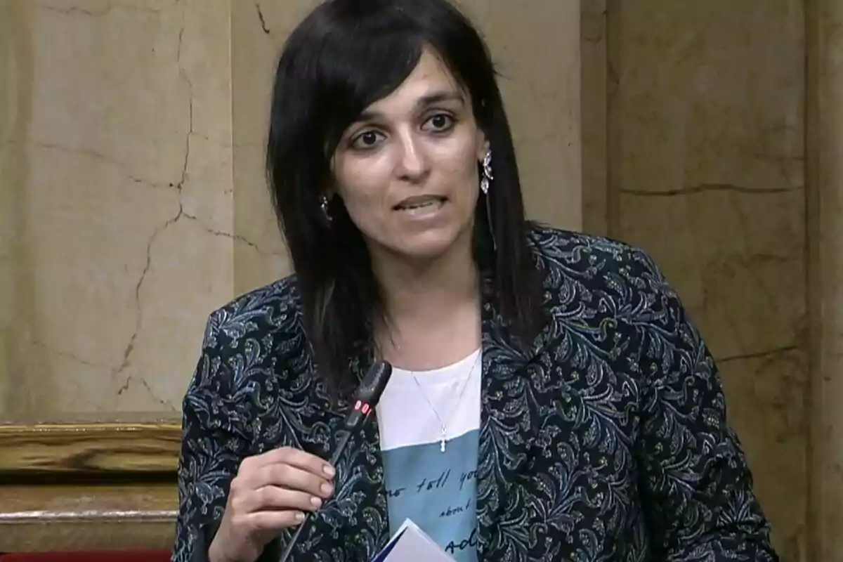 Dark-haired woman speaking into a microphone in a formal setting with marble walls.