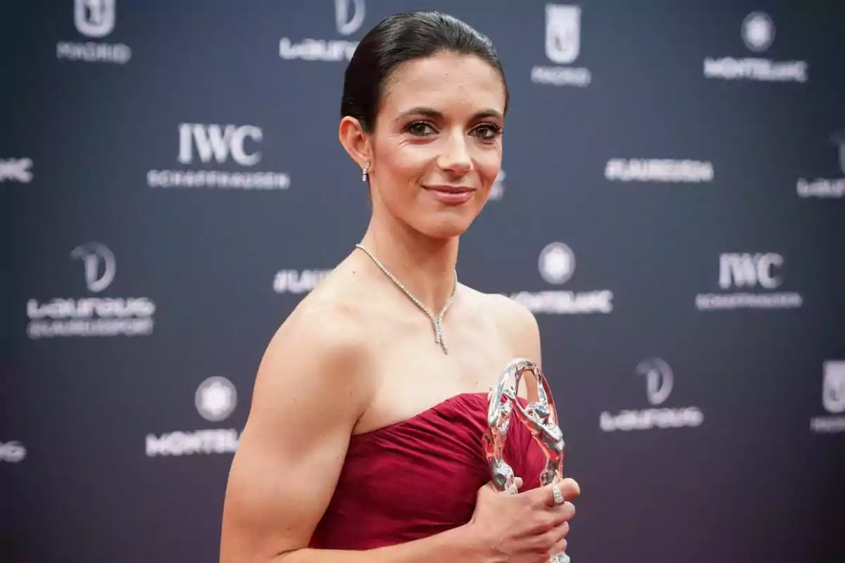 Dona amb vestit vermell elegant sosté un trofeu platejat mentre somriu en una catifa vermella amb un fons de logotips de patrocinadors.