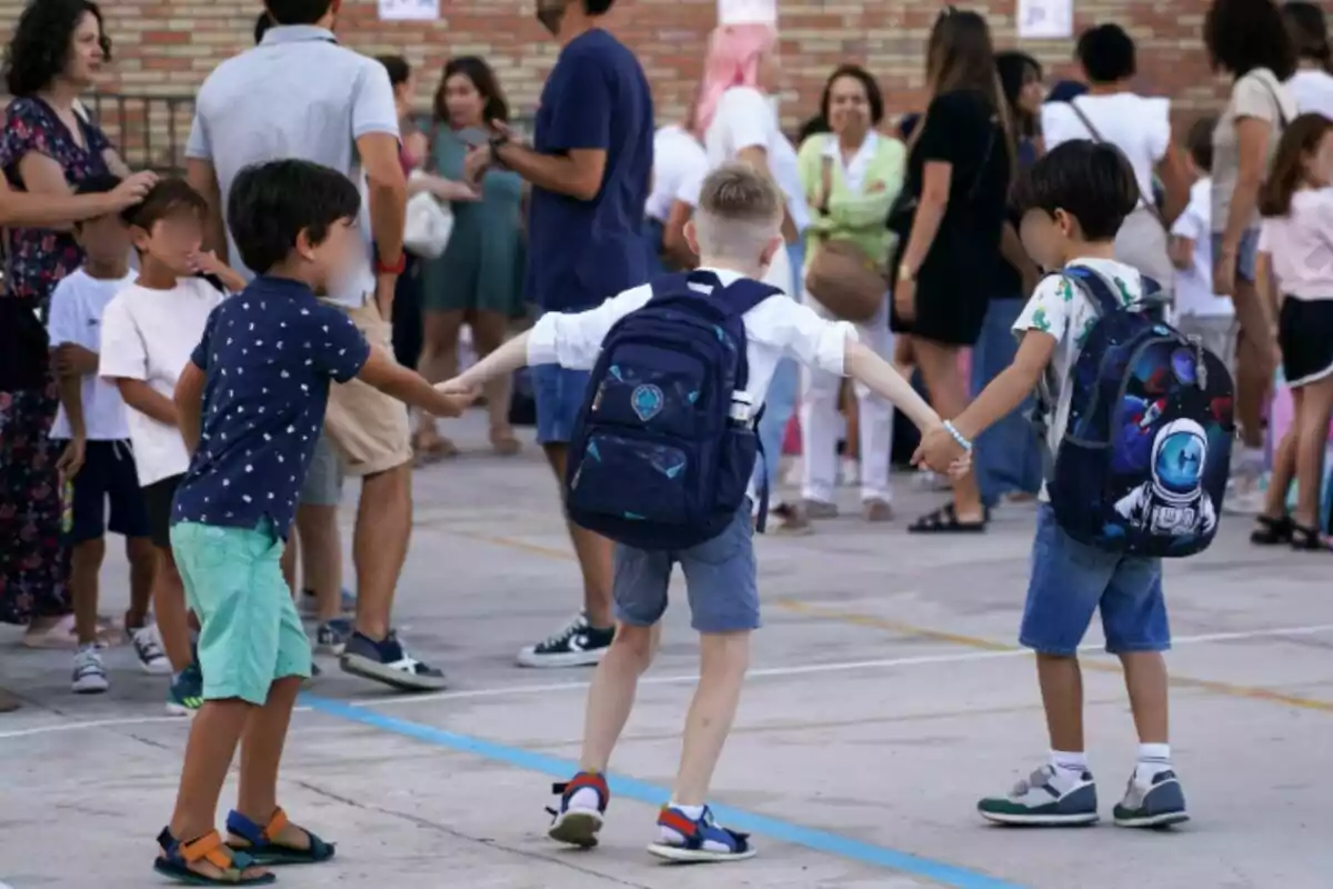 Tres nens amb motxilles es donen les mans en un pati escolar mentre diversos adults i altres nens són al fons. Tres nens amb motxilles es donen les mans en un pati escolar mentre diversos adults i altres nens són al fons.
