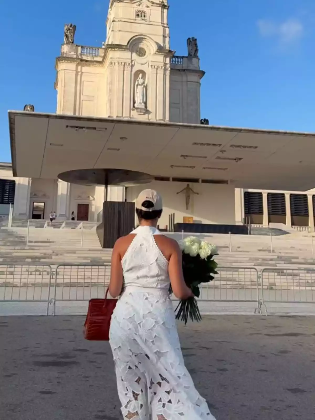 Dona vestida de blanc amb un ram de flors i una bossa vermella caminant cap a una església gran sota un cel blau Dona vestida de blanc amb un ram de flors i una bossa vermella caminant cap a una església gran sota un cel blau
