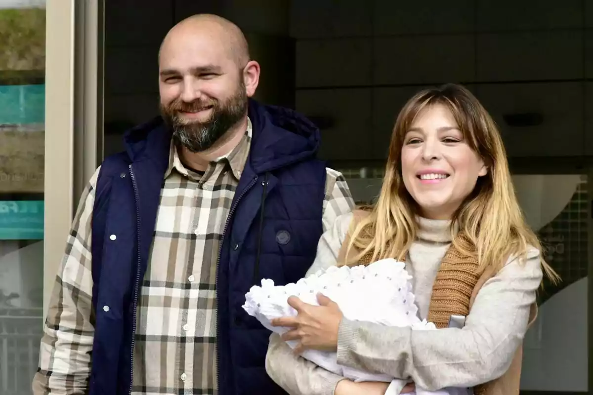 Una parella somrient posa davant d’un edifici mentre la dona sosté en braços un nadó embolcallat amb una manta blanca. Una parella somrient posa davant d’un edifici mentre la dona sosté en braços un nadó embolcallat amb una manta blanca.