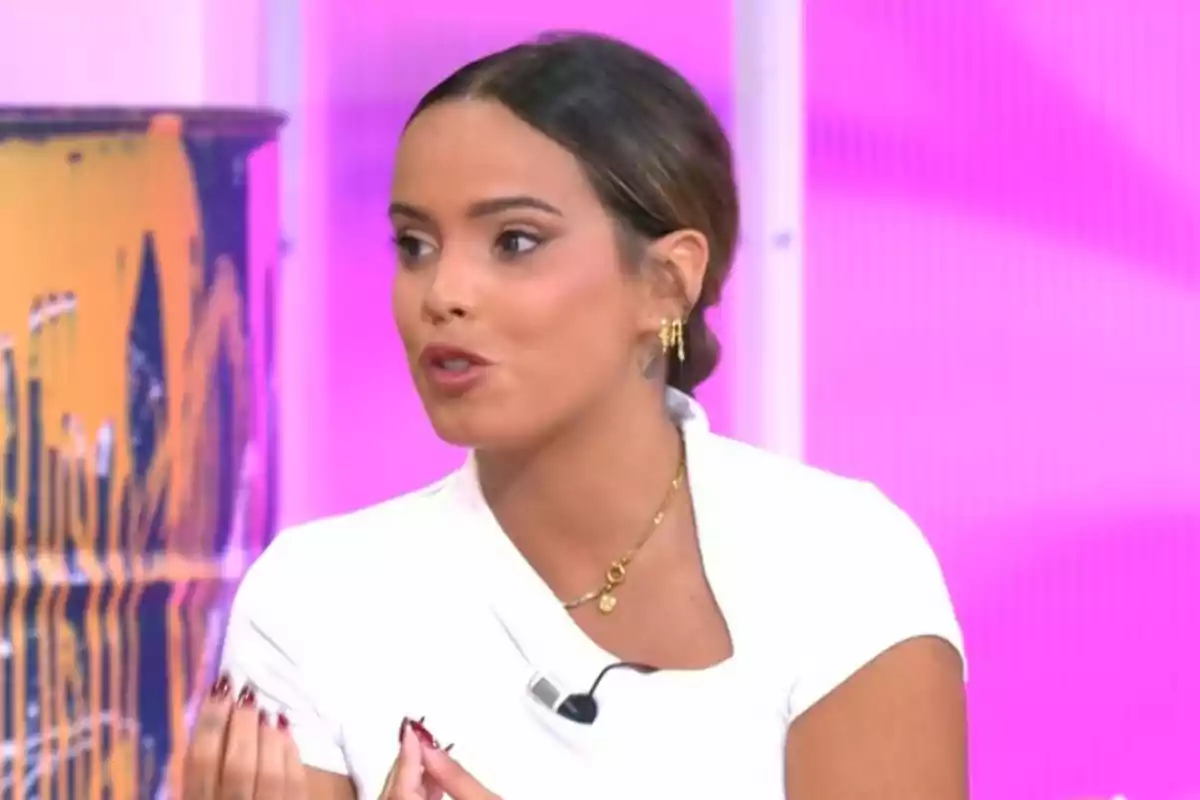Dona jove amb els cabells recollits i vestida de blanc parlant en un plató de televisió amb fons rosa Dona jove amb els cabells recollits i vestida de blanc parlant en un plató de televisió amb fons rosa