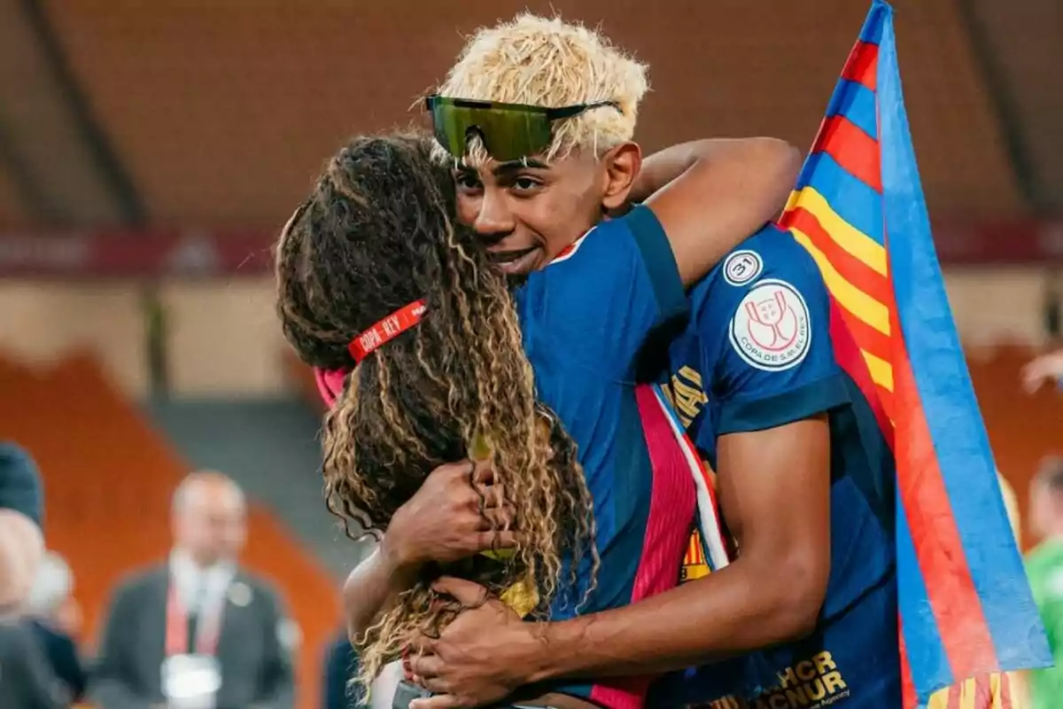 Jugador de fútbol con cabello rubio y gafas oscuras abraza a una persona mientras sostiene una bandera colorida en un estadio Jugador de fútbol con cabello rubio y gafas oscuras abraza a una persona mientras sostiene una bandera colorida en un estadio