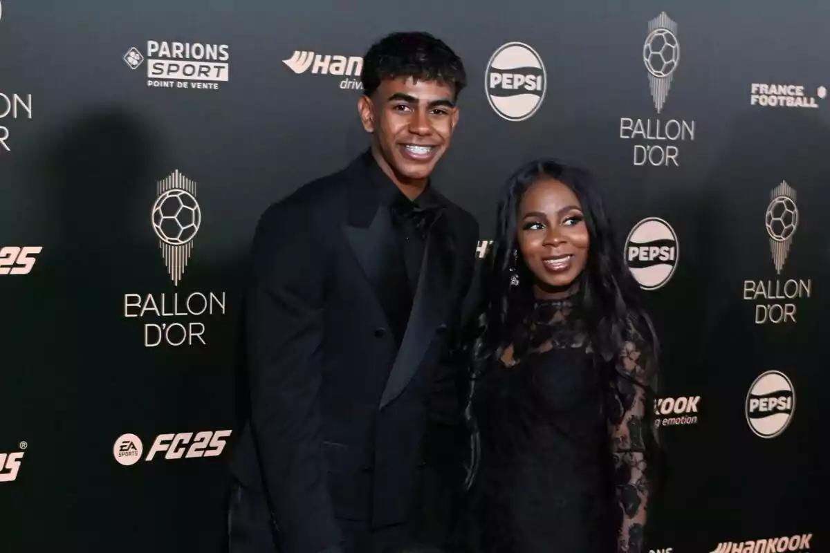 Un joven y una mujer posan sonrientes en la alfombra roja de la gala del Balón de Oro, ambos vestidos de negro elegante, con un fondo decorado con logotipos del evento y patrocinadores. Un joven y una mujer posan sonrientes en la alfombra roja de la gala del Balón de Oro, ambos vestidos de negro elegante, con un fondo decorado con logotipos del evento y patrocinadores.