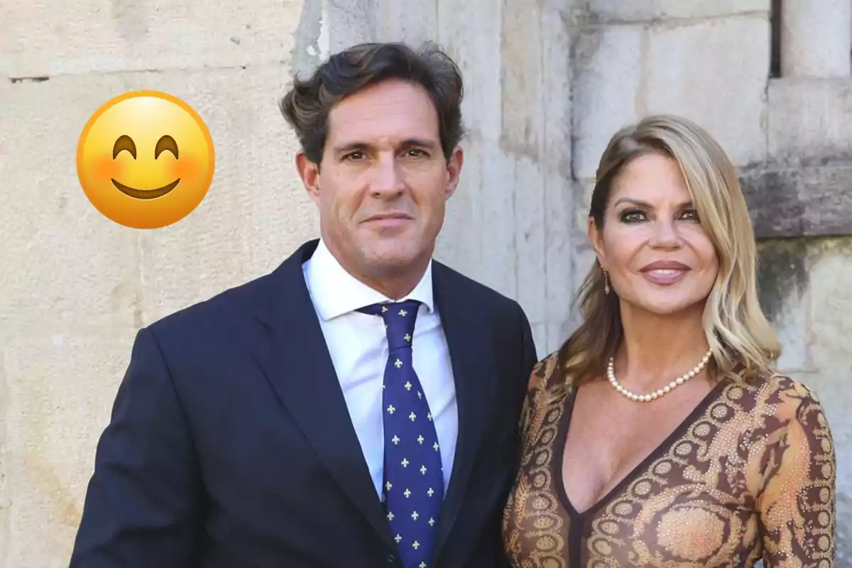 Parella elegantament vestida posant davant d’una paret de pedra amb un gran emoji somrient a l’esquerra