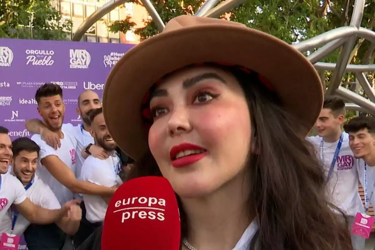 Una dona amb barret i maquillatge cridaner és entrevistada en un esdeveniment a l’aire lliure amb un grup de persones somrients al fons i un micròfon vermell d’Europa Press en primer pla. Una dona amb barret i maquillatge cridaner és entrevistada en un esdeveniment a l’aire lliure amb un grup de persones somrients al fons i un micròfon vermell d’Europa Press en primer pla.