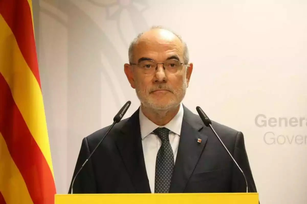 Older man with glasses and a gray beard speaking at a press conference with microphones and a Catalan flag beside him Older man with glasses and a gray beard speaking at a press conference with microphones and a Catalan flag beside him