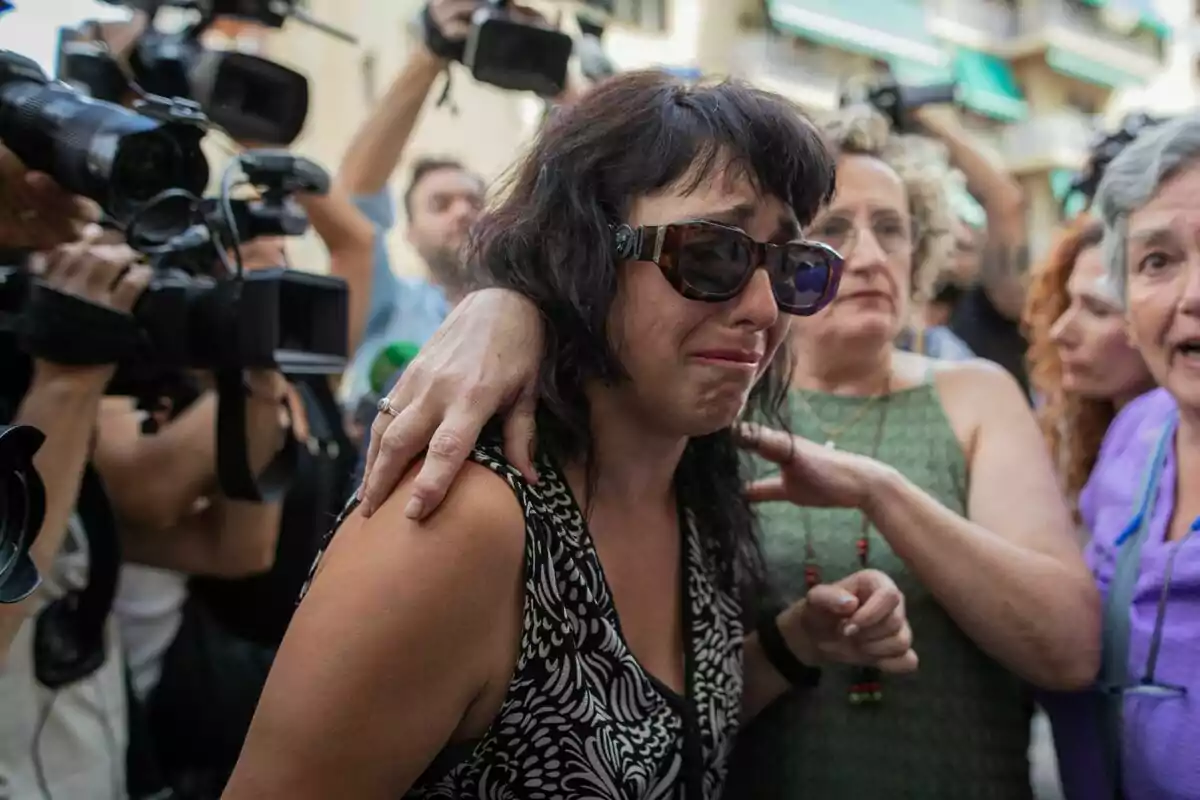 Dona plorant amb ulleres de sol envoltada de persones i càmeres de premsa en un ambient de tensió i suport Dona plorant amb ulleres de sol envoltada de persones i càmeres de premsa en un ambient de tensió i suport