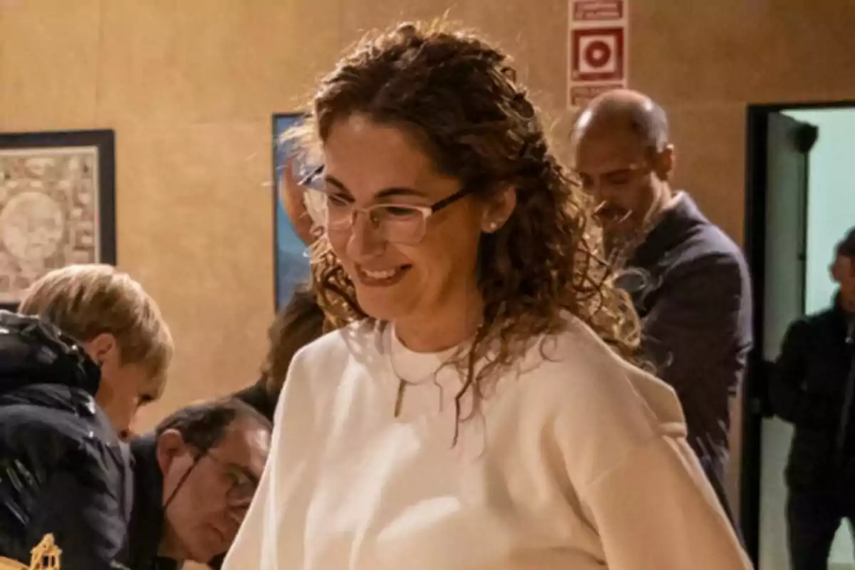 Dona de cabells arrissats i ulleres somriu mentre participa en una activitat en un espai interior amb diverses persones al fons. Dona de cabells arrissats i ulleres somriu mentre participa en una activitat en un espai interior amb diverses persones al fons.