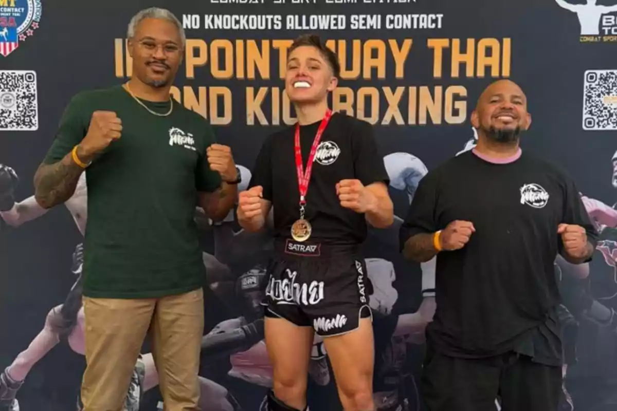 Tres persones posen somrients amb actitud de victòria davant d’un cartell de Muay Thai i Kickboxing, la persona del centre porta una medalla i protector bucal. Tres persones posen somrients amb actitud de victòria davant d’un cartell de Muay Thai i Kickboxing, la persona del centre porta una medalla i protector bucal.