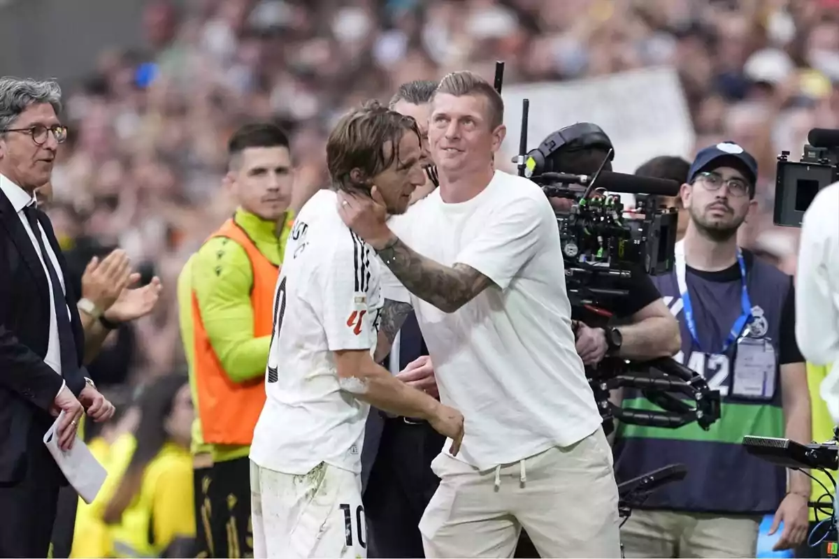 Un futbolista amb uniforme blanc és abraçat per un home vestit de blanc mentre diverses persones observen i una càmera professional grava l’escena en un estadi ple de públic. Un futbolista amb uniforme blanc és abraçat per un home vestit de blanc mentre diverses persones observen i una càmera professional grava l’escena en un estadi ple de públic.