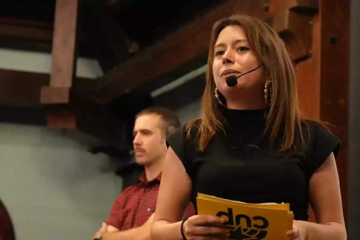 A woman with a headset microphone holds some yellow papers while speaking; in the background, a man in a red jersey with an earpiece watches alertly.