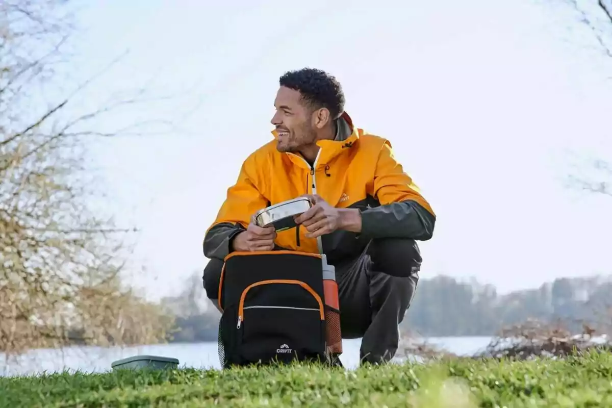 Man in an orange jacket sitting on the grass by a lake holding a food container and a black backpack Man in an orange jacket sitting on the grass by a lake holding a food container and a black backpack