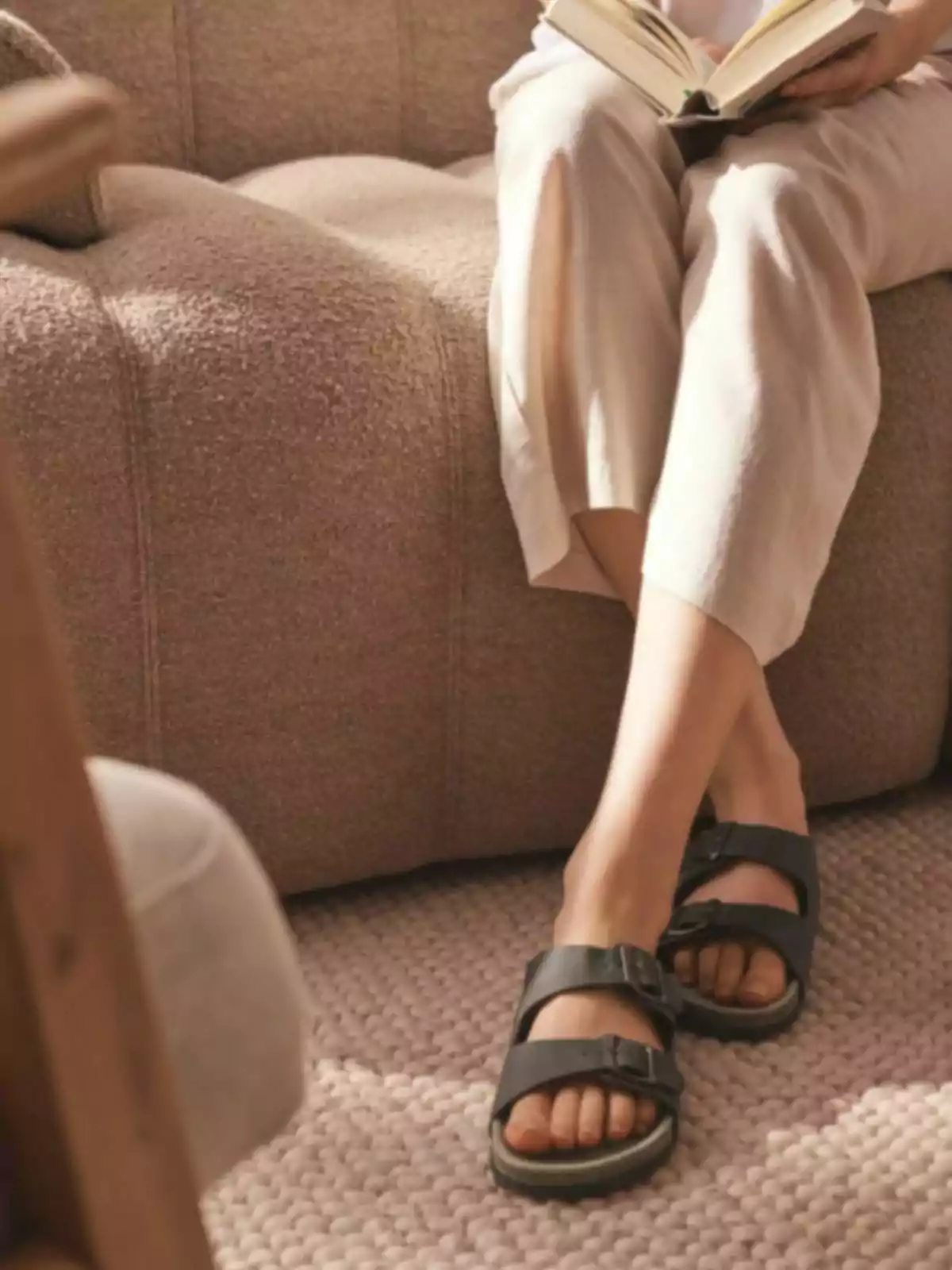 Person sitting on a beige armchair reading a book, wearing light-colored pants and black sandals on a woven rug. Person sitting on a beige armchair reading a book, wearing light-colored pants and black sandals on a woven rug.