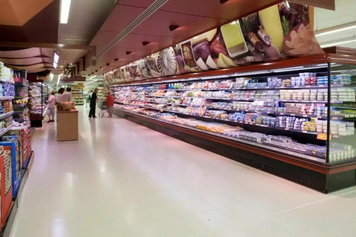 Passadís de supermercat amb vitrines refrigerades plenes de productes i diverses persones fent la compra Passadís de supermercat amb vitrines refrigerades plenes de productes i diverses persones fent la compra
