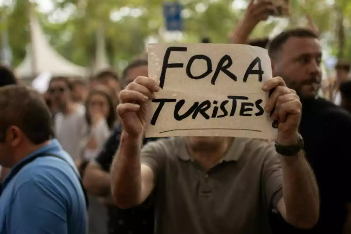 Persona sosté un cartell amb el missatge Fora Turistes en una manifestació envoltada de més gent. Persona sosté un cartell amb el missatge Fora Turistes en una manifestació envoltada de més gent.