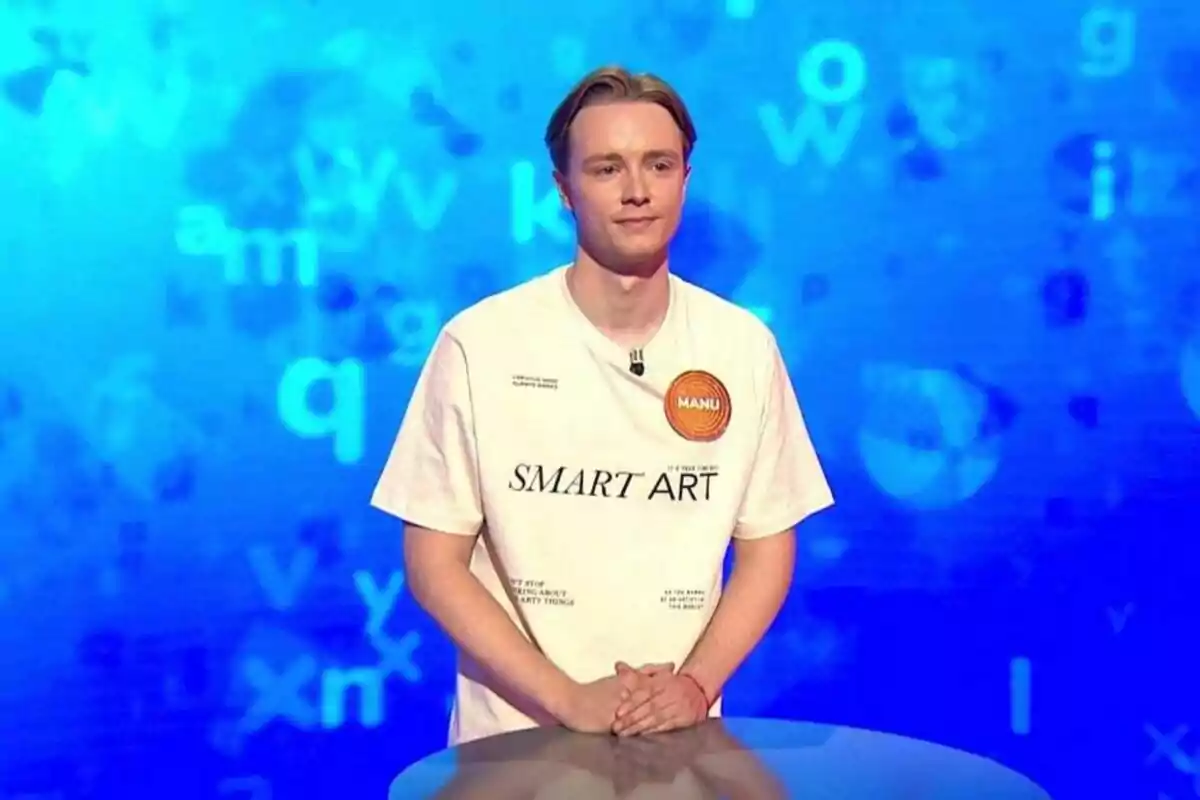 Manu Pascual, standing behind a lectern on a television set with a blue background and scattered letters, is wearing a white t-jersey with the text SMART ART and an orange circle that says MANU. Manu Pascual, standing behind a lectern on a television set with a blue background and scattered letters, is wearing a white t-jersey with the text SMART ART and an orange circle that says MANU.