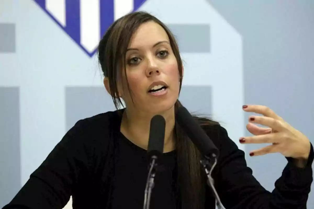 Young woman speaking in front of two microphones with a light blue background and a geometric logo behind her