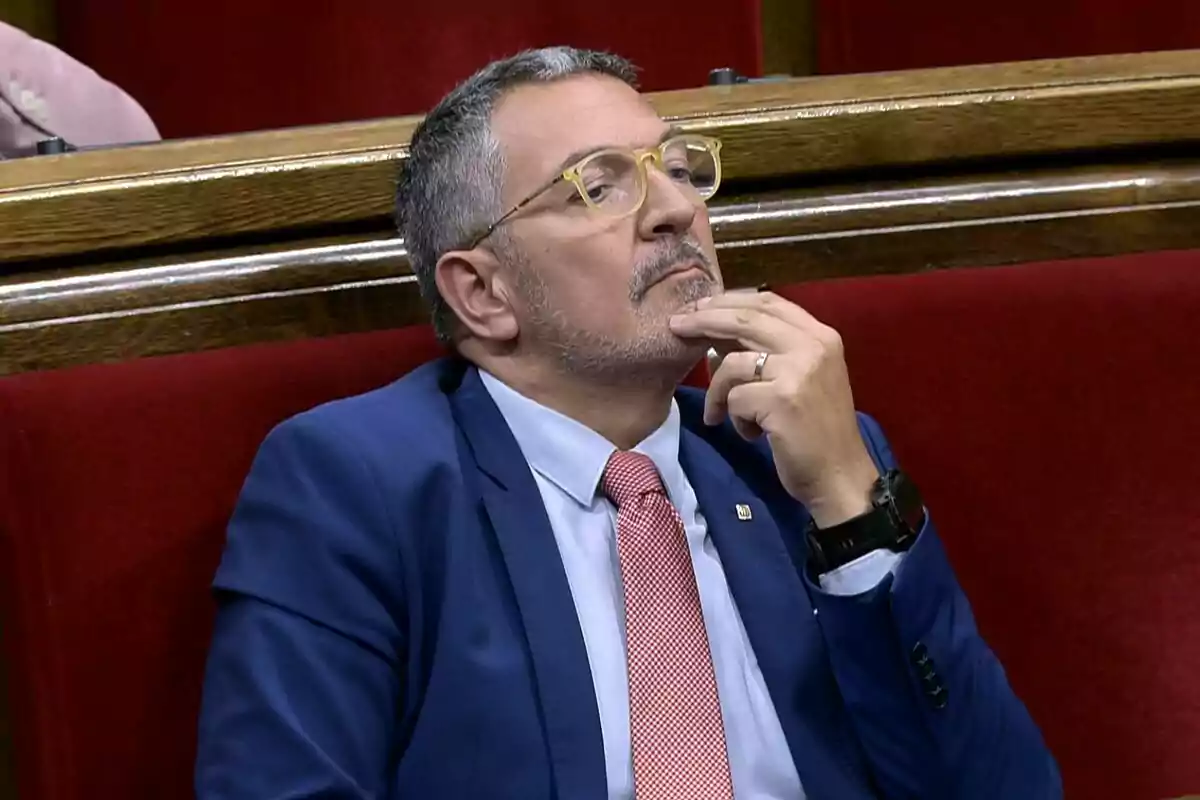 Man with glasses and a blue suit sitting on a red bench with a thoughtful expression and his hand on his chin