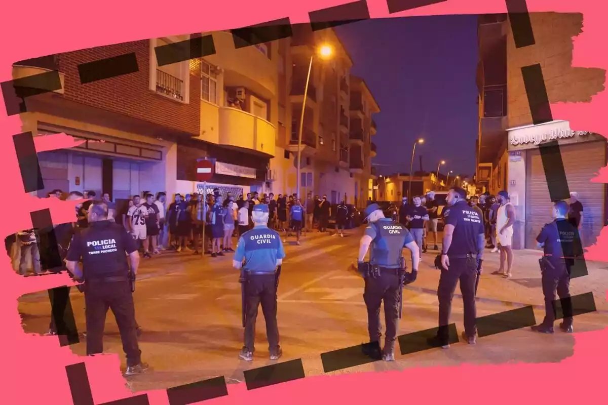 Un grup de policies i guàrdies civils formen una barrera davant d’un grup de persones en un carrer de nit, amb edificis residencials i llums enceses, tot emmarcat per una vora rosa amb línies negres.