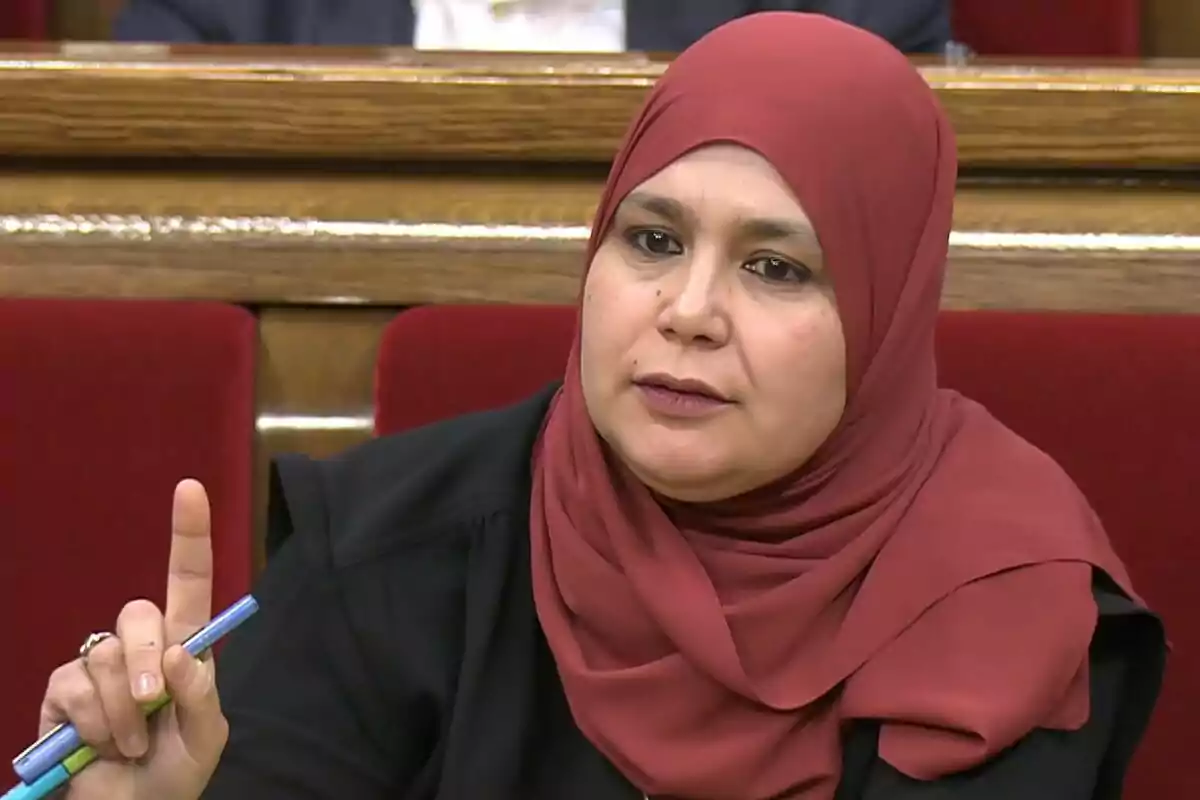 Dona amb hijab vermell asseguda en una sala amb seients vermells aixecant un dit i sostenint bolígrafs