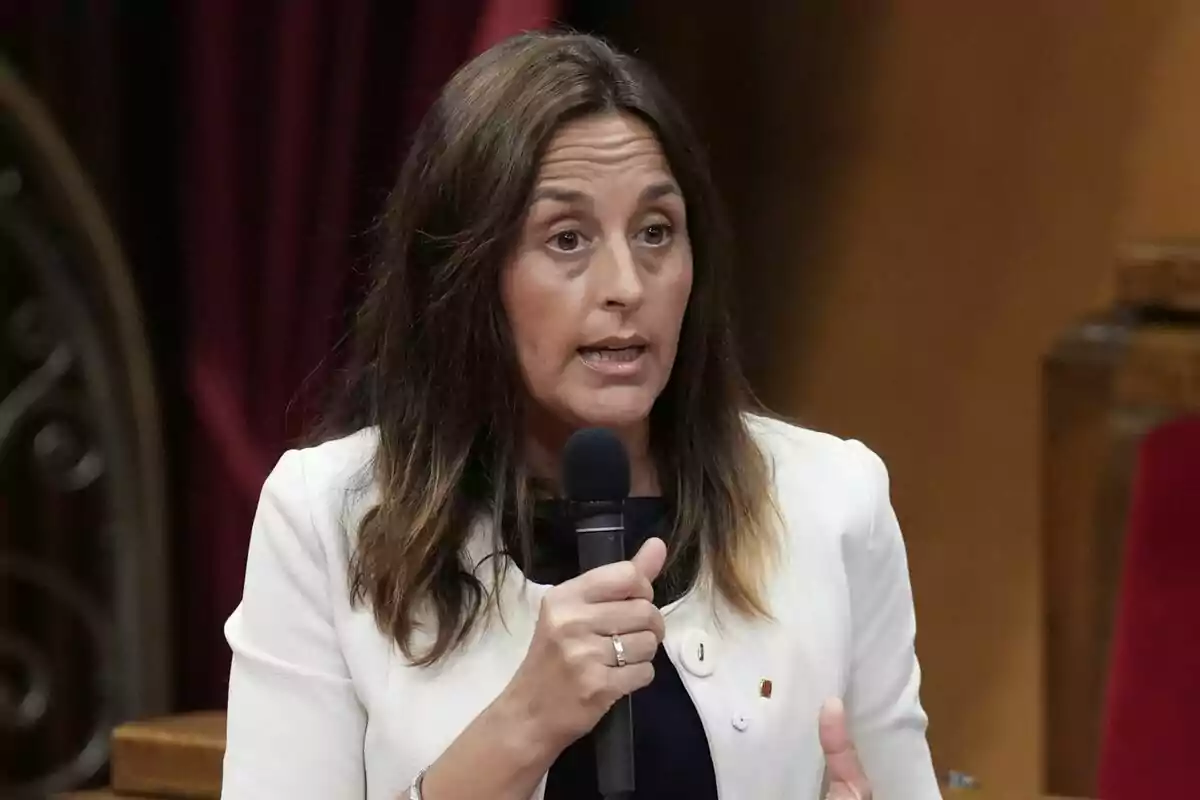 Brown-haired woman speaking in public while holding a microphone and wearing a white jacket Brown-haired woman speaking in public while holding a microphone and wearing a white jacket