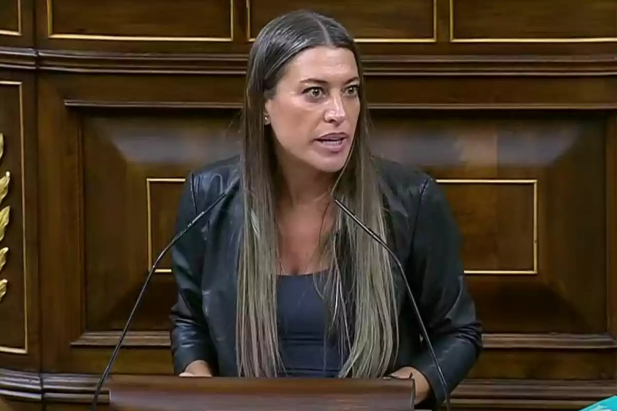 Woman with long, straight hair speaking at a wooden podium with a background of wooden panels Woman with long, straight hair speaking at a wooden podium with a background of wooden panels